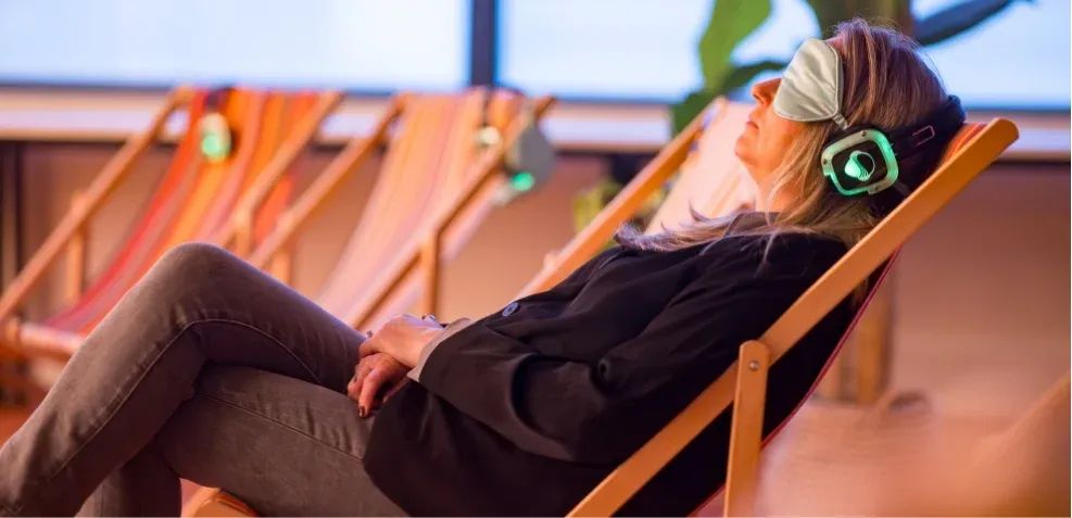 A woman in an eye mask and glowing headphones relaxes in a wooden deck chair.