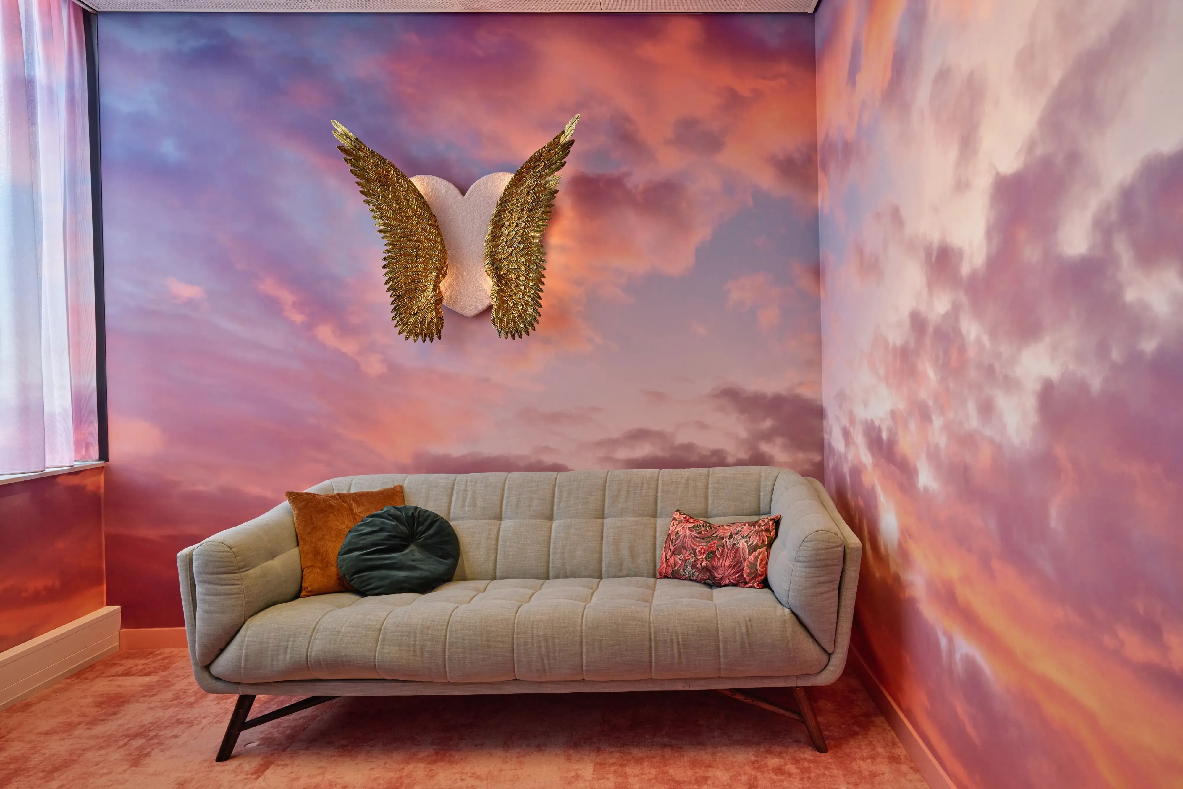 Lichte tweezitsbank tegen een muur met roze en paarse wolkenprint, met daarboven gouden vleugels in de vorm van een hart; op de bank liggen sierkussens in warme tinten.