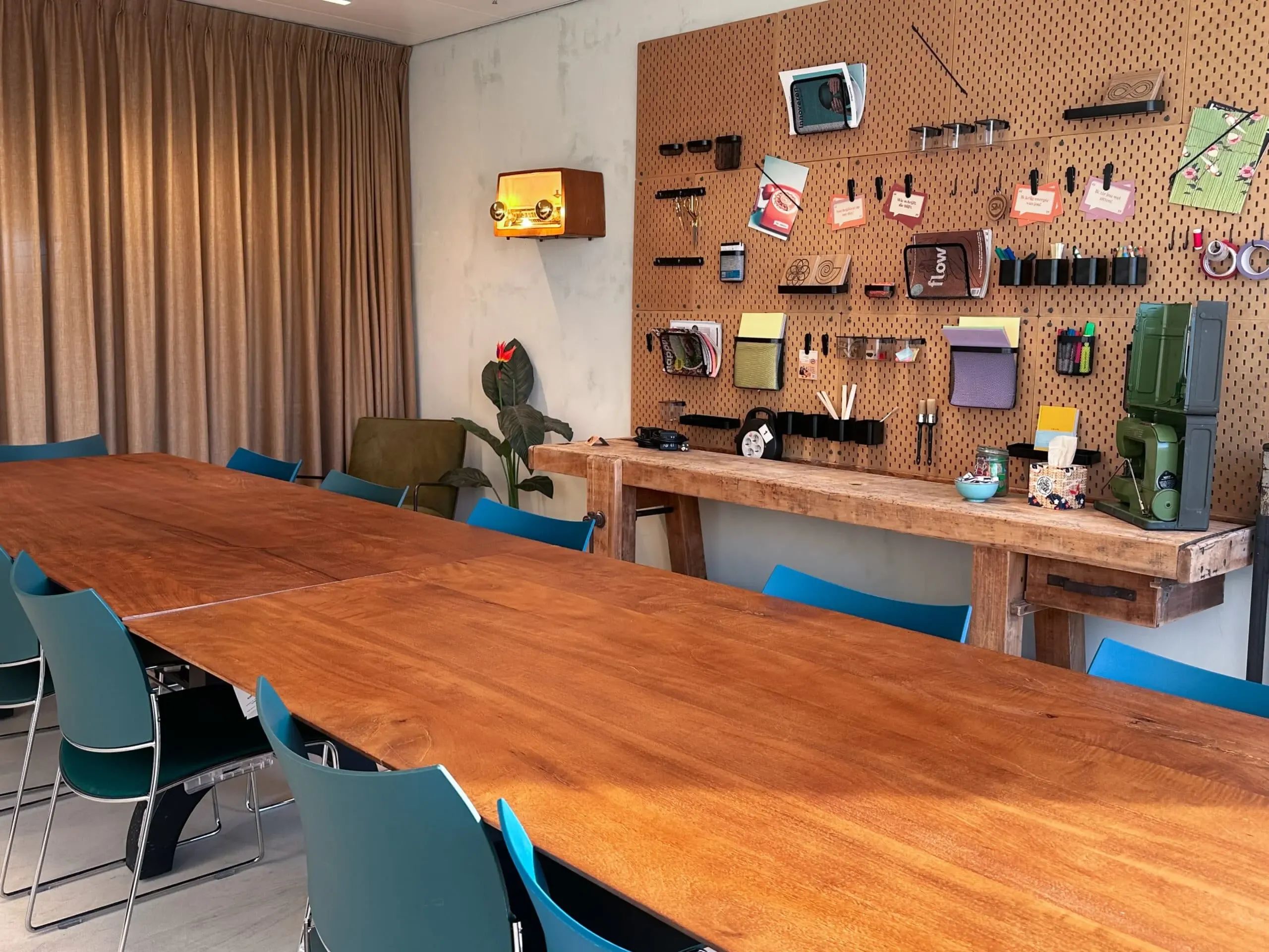 Creatieve vergaderruimte met lange houten tafel, blauwe stoelen en pegboard wand met kantooraccessoires in moderne kantooromgeving.