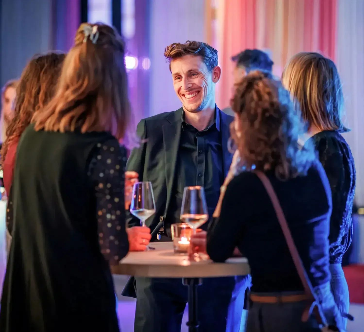 Young man smiling and conversing with friends at a vibrant social gathering with drinks.