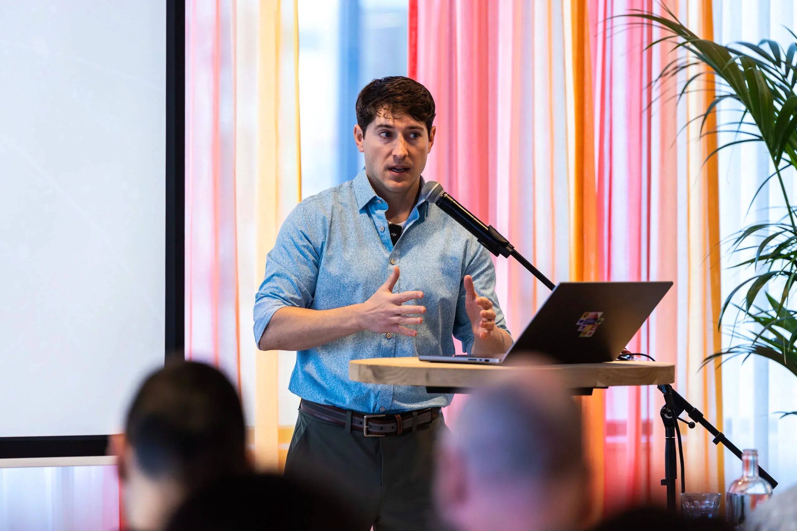 Een man in een blauw shirt spreekt tijdens een presentatie vanaf een podium met een microfoon en laptop.