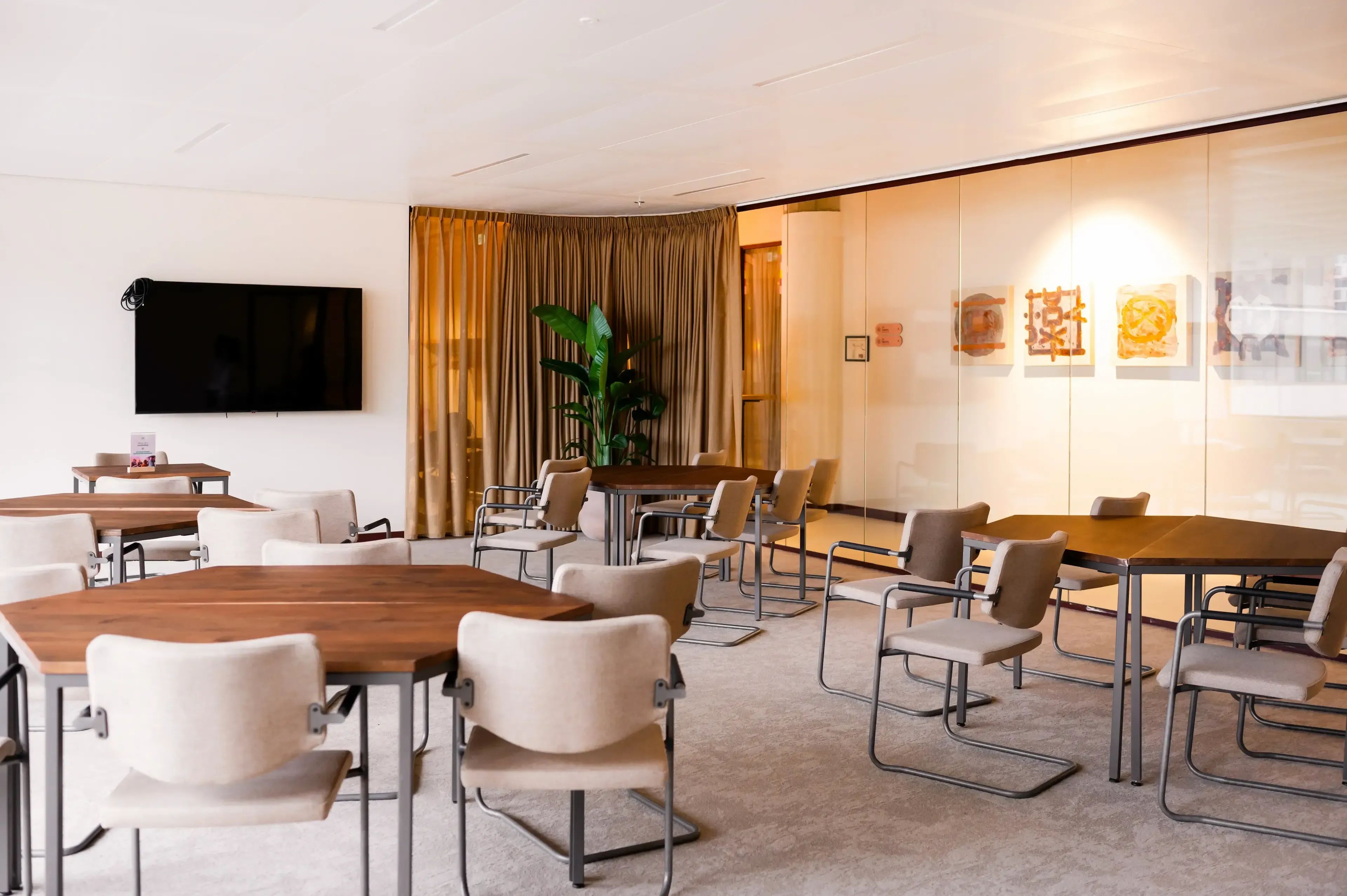 Moderne vergaderruimte met meerdere houten tafels, stoelen en een grote tv aan de muur.