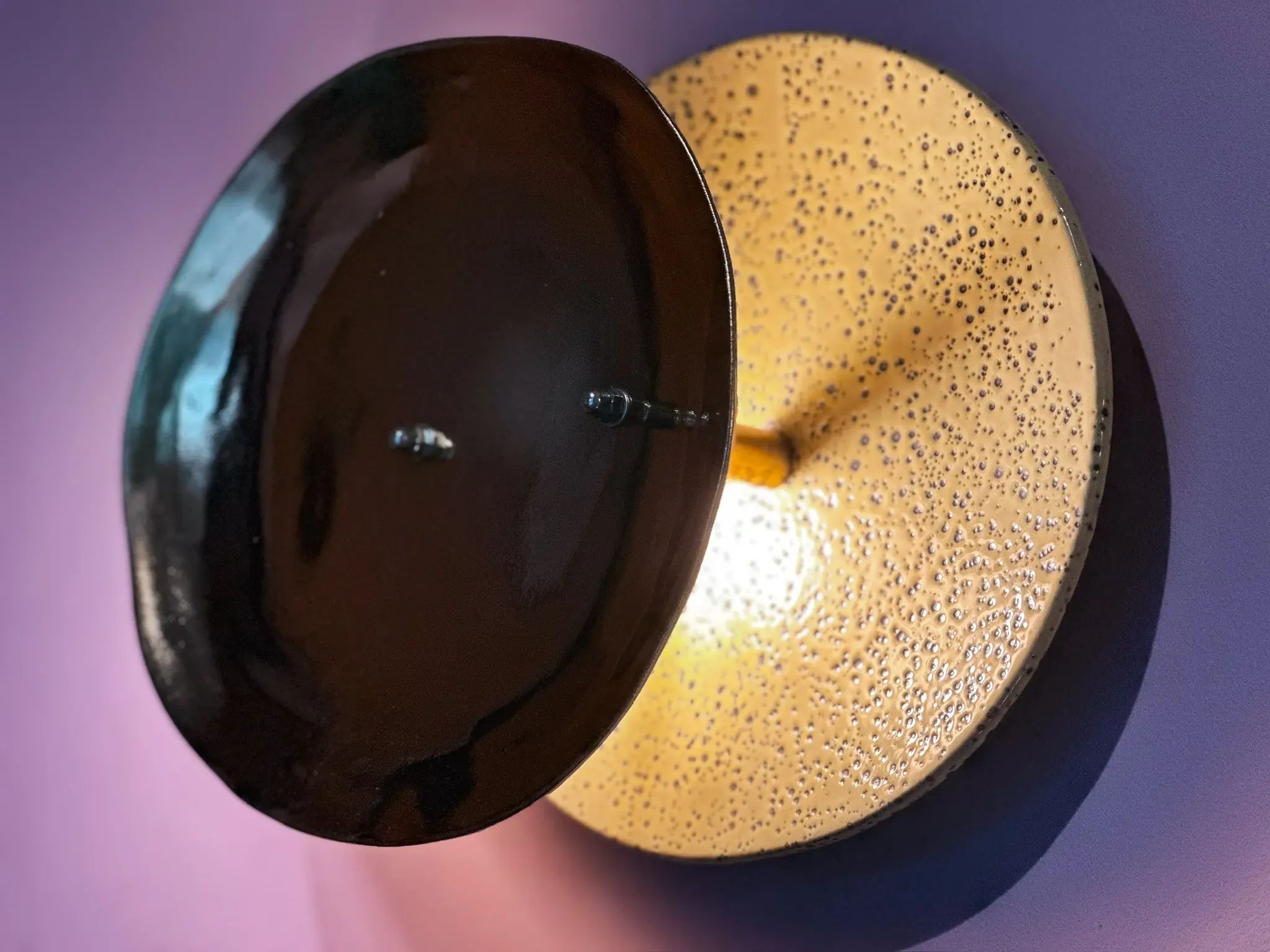 Een moderne wandlamp met overlappende donkere en gestructureerde lichtgekleurde schijven die een paarse muur verlichten.