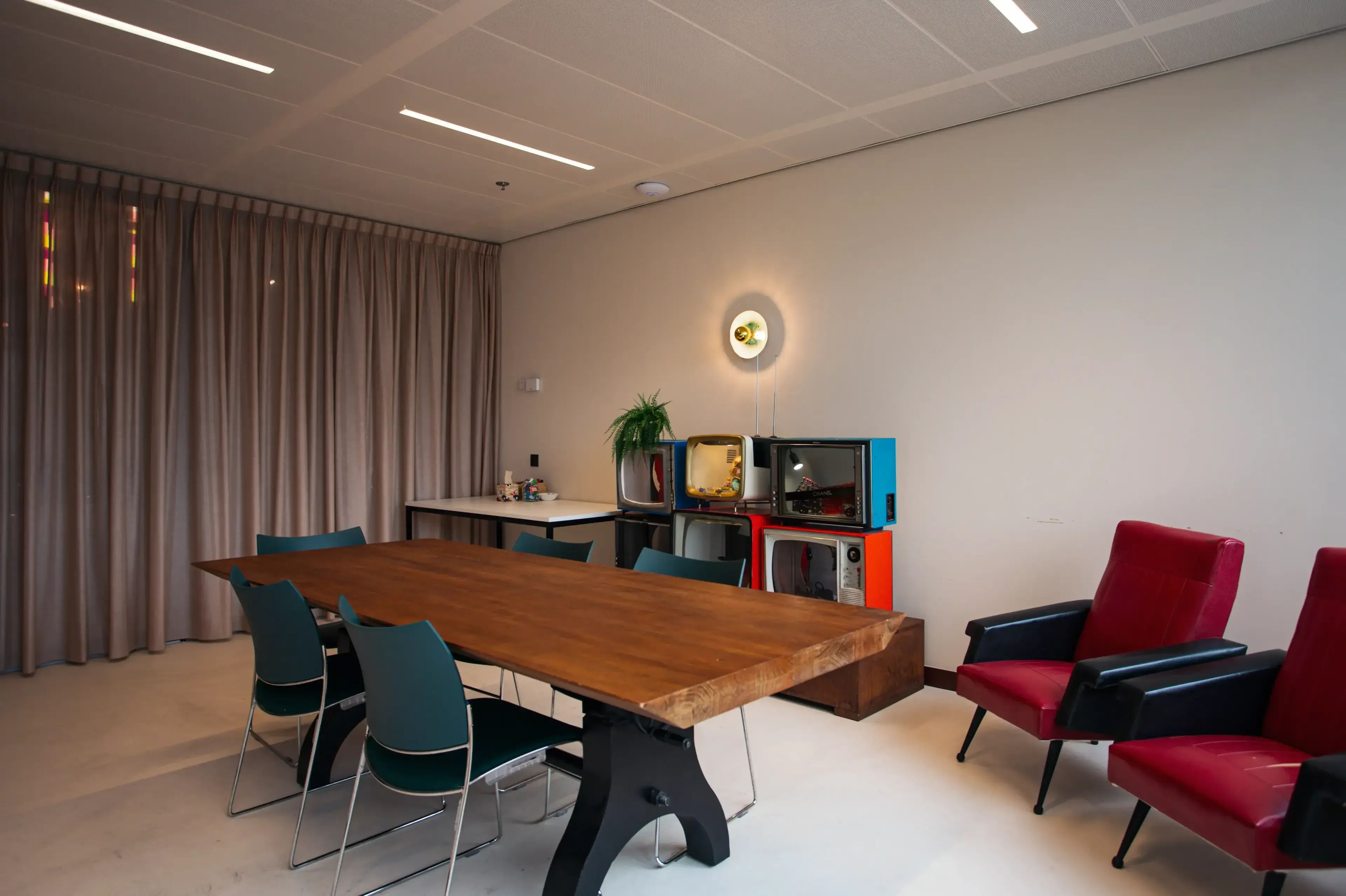 Moderne vergaderruimte met een grote houten tafel, rode fauteuils en gestapelde vintage televisies.