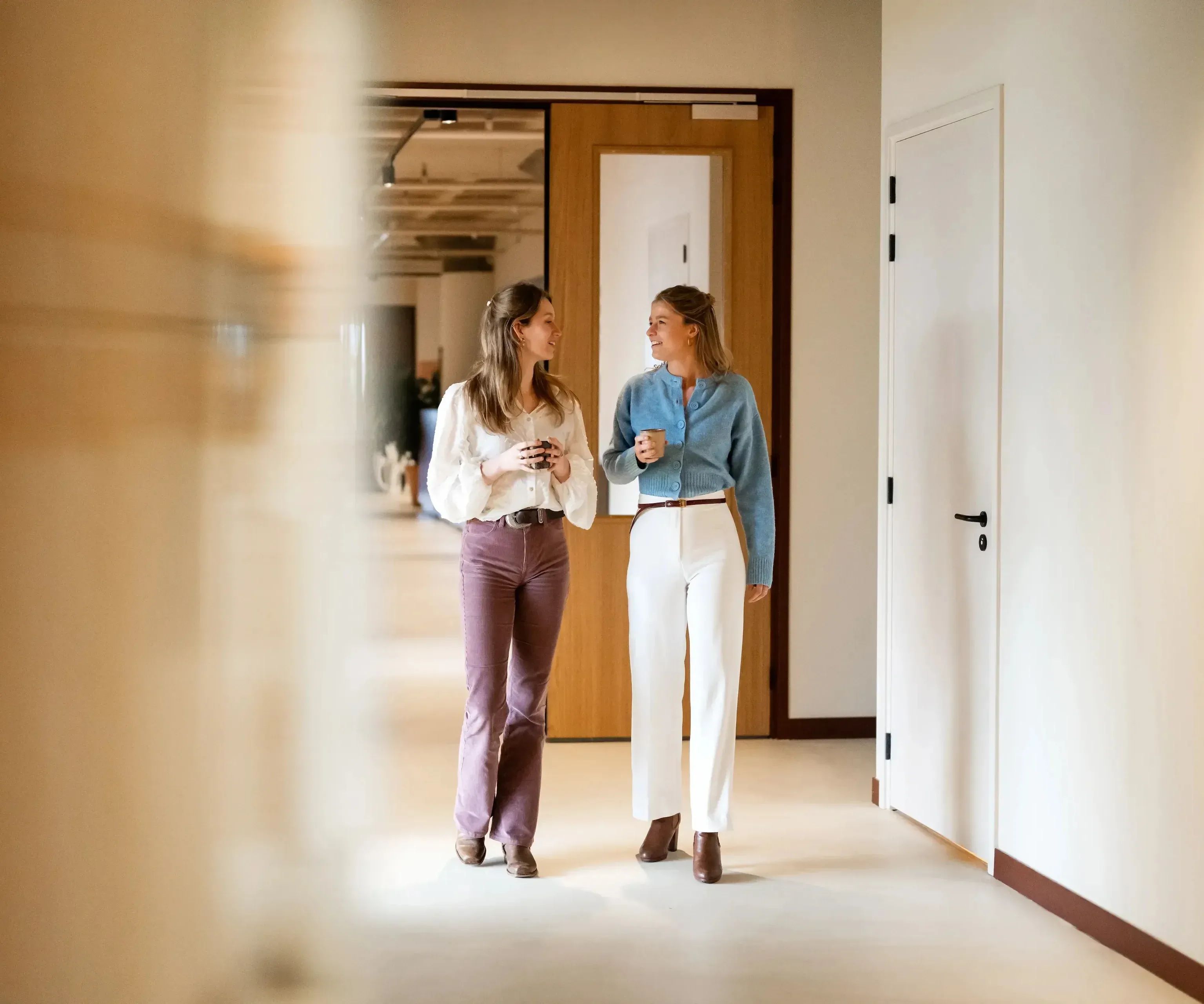 Twee vrouwen lopen en praten in een lichte, moderne kantoorgang.