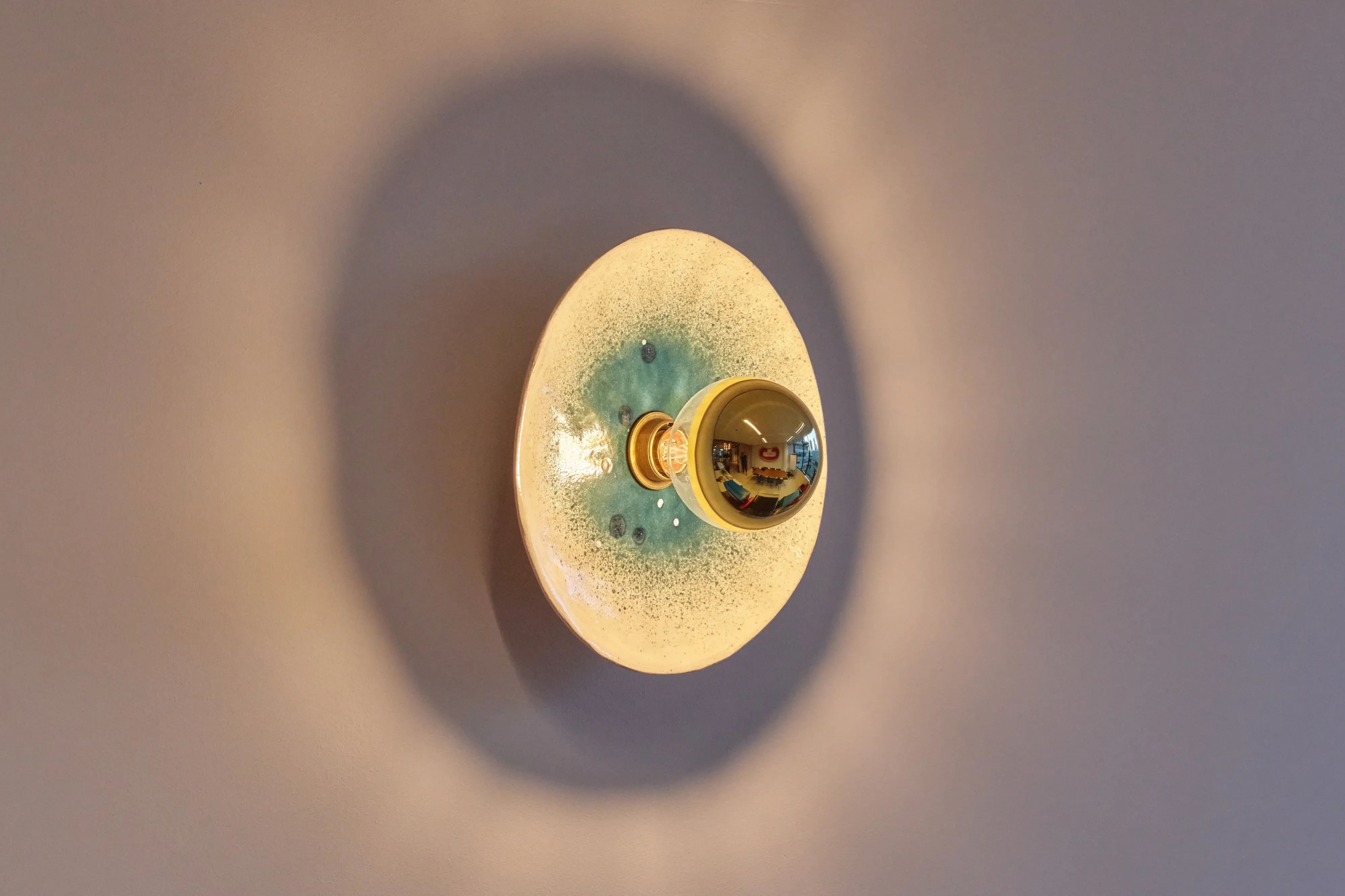 Organisch gevormde wandlamp met gespikkelde witte en blauwgroene kap, die warm licht verspreidt op een muur.