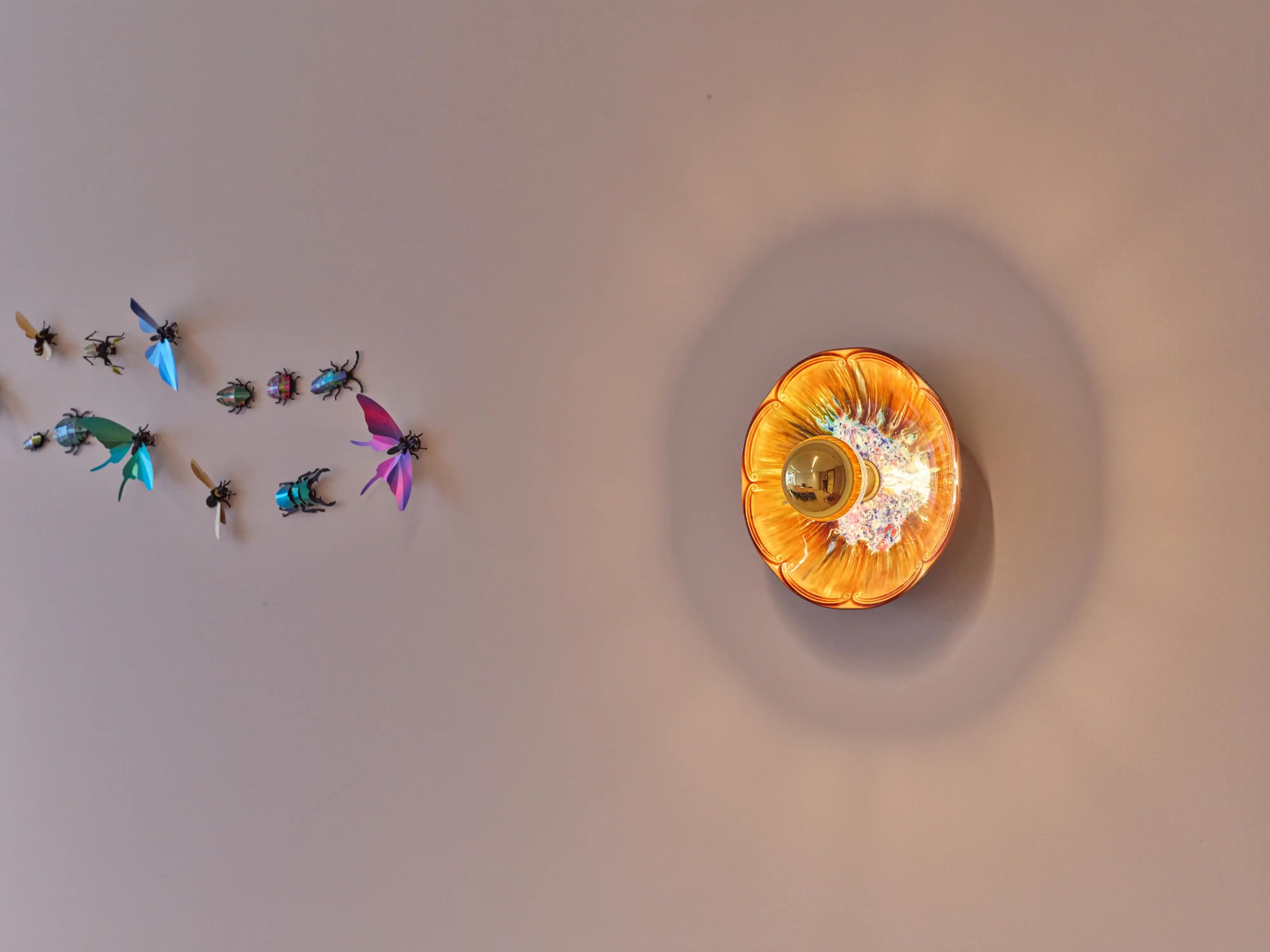 Op een beige muur is een gebogen reeks kleurrijke insectachtige figuren en een cirkelvormig oranje licht te zien.