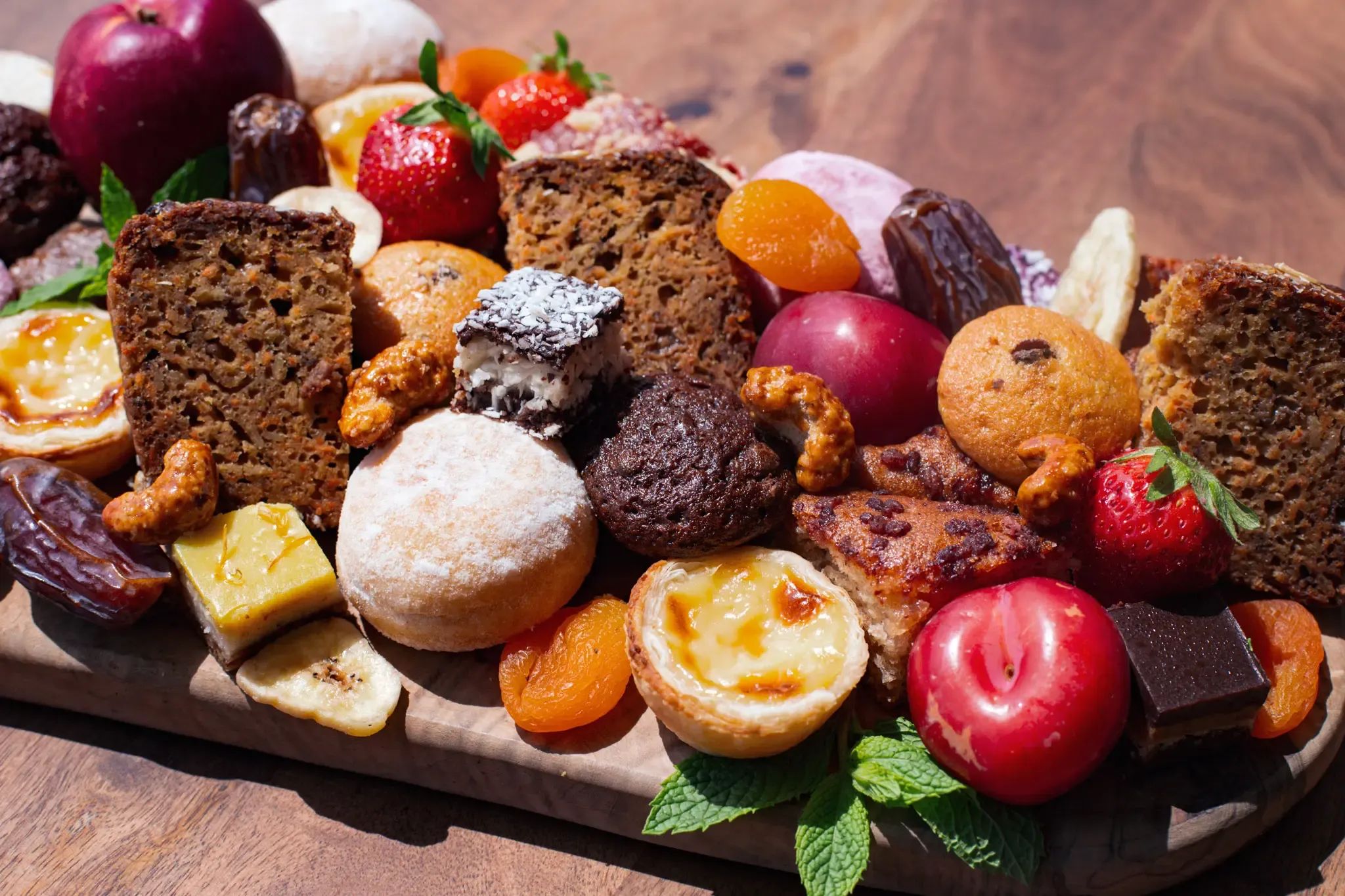 Assorted pastries, cakes, fresh and dried fruits, and chocolate on a wooden board.