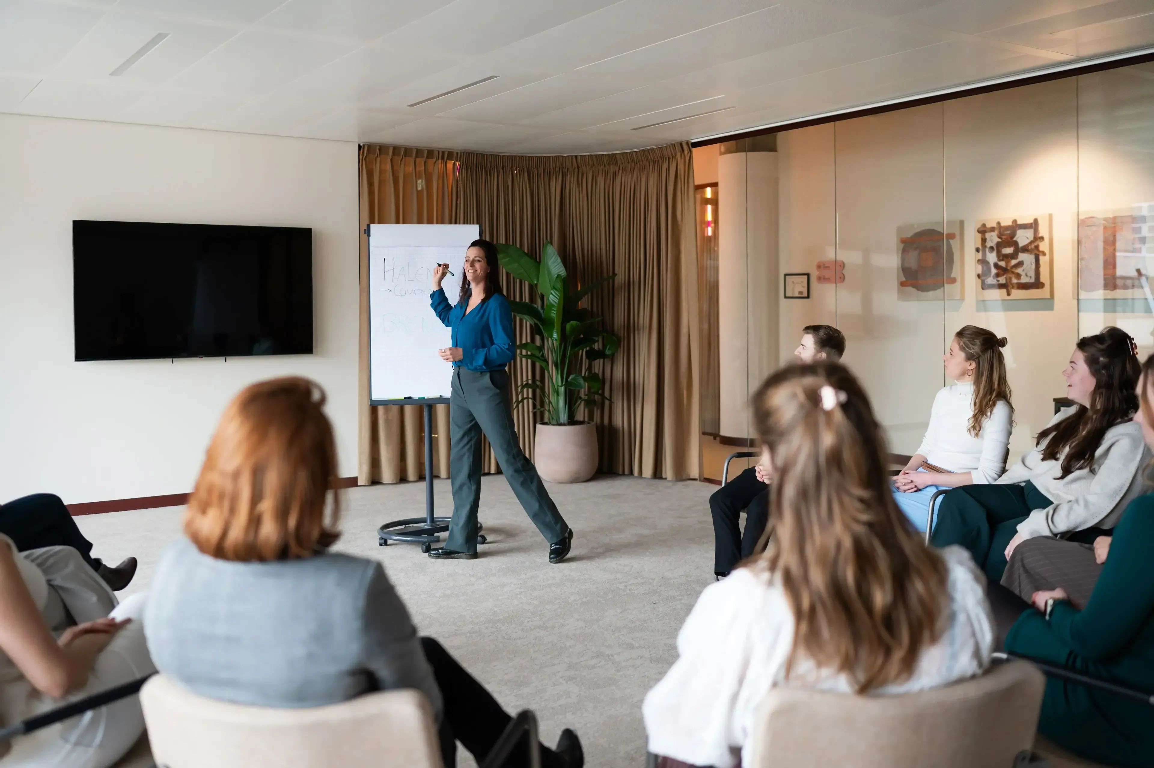 Vrouw in blauw shirt geeft een presentatie aan een groep mensen die in een lichte, moderne vergaderruimte zitten.