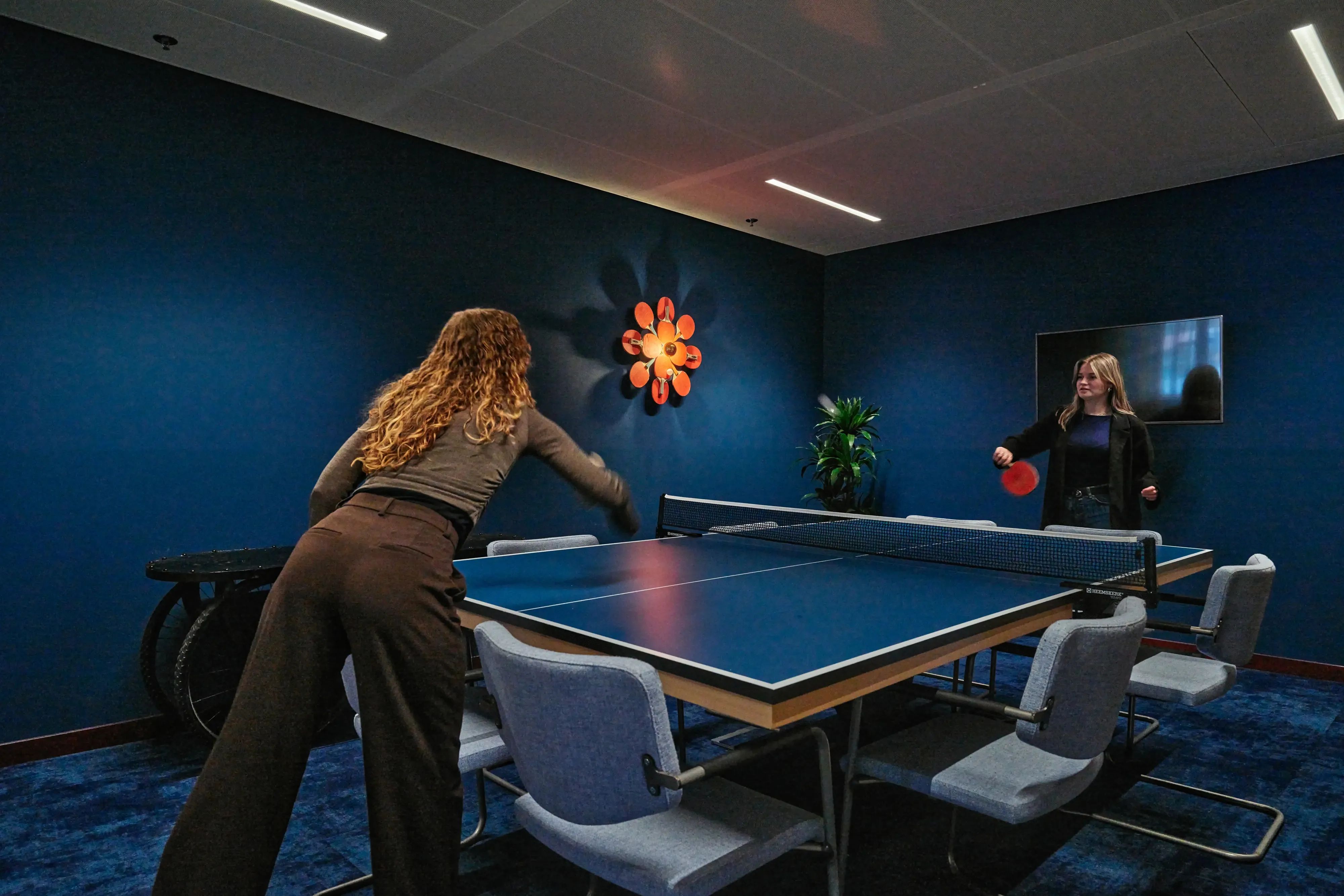 Twee vrouwen spelen tafeltennis in een moderne blauwe kamer met een unieke stervormige lamp.