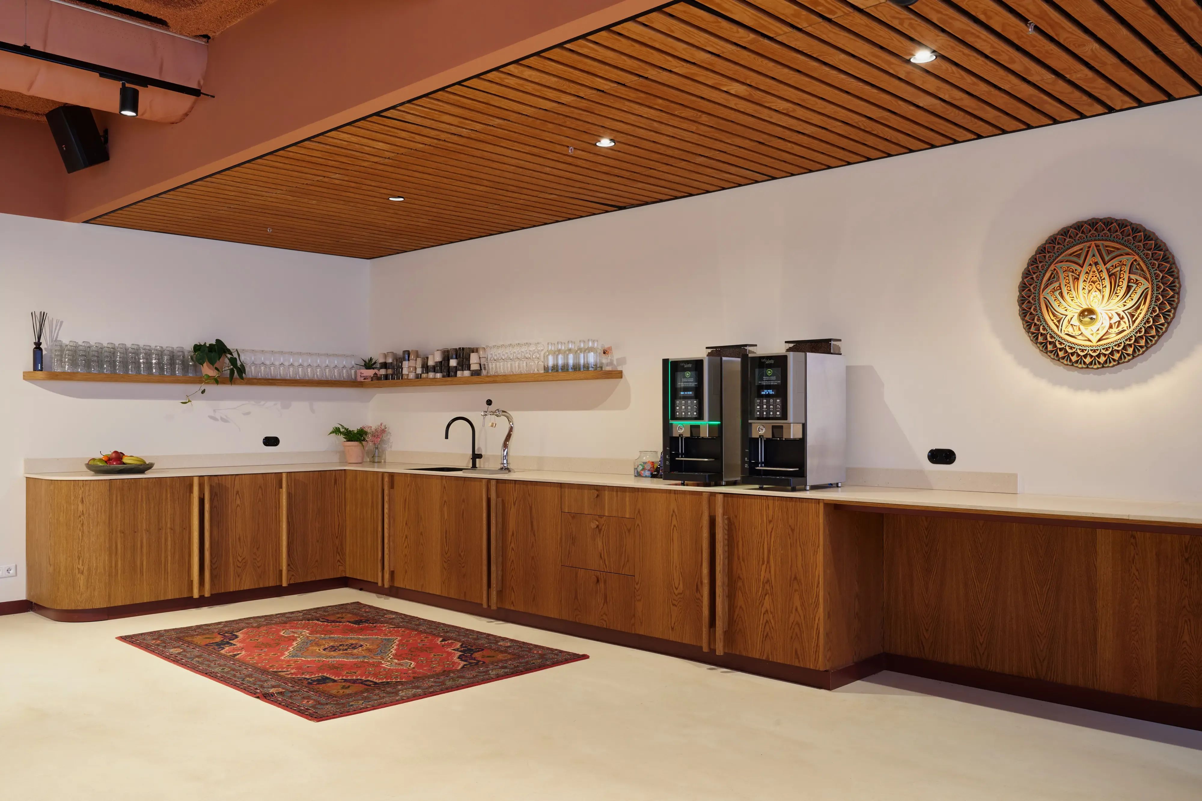 Moderne bar met houten kasten, twee koffiemachines, een gootsteen en een decoratief tapijt.