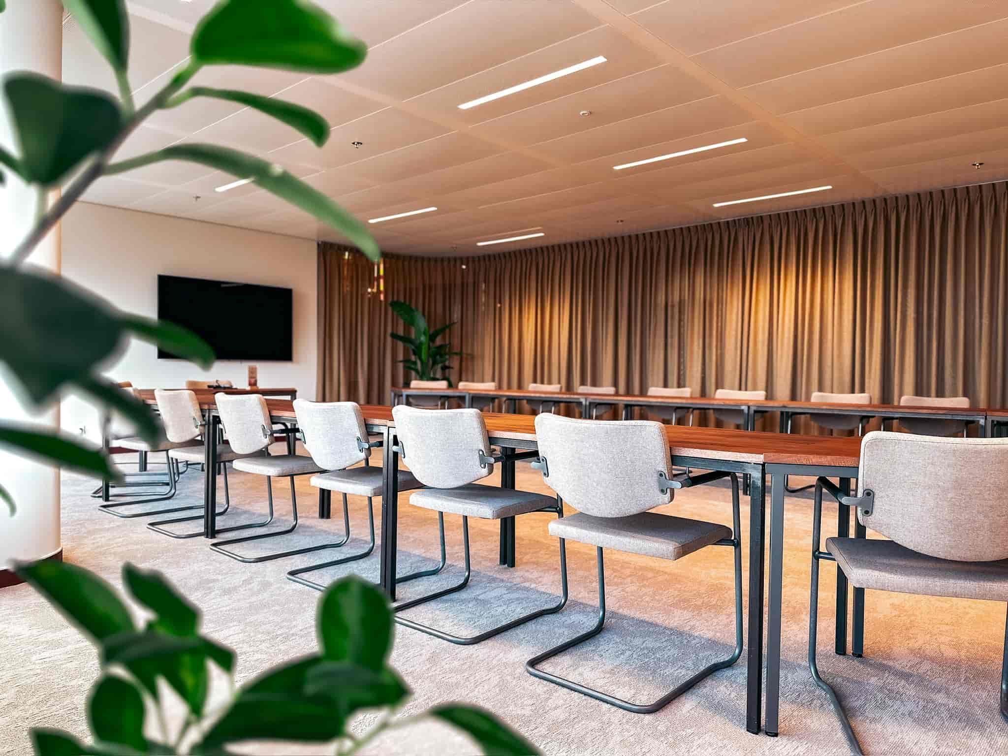 Moderne, lege vergaderzaal met een lange tafel, stoelen, groot scherm en natuurlijk licht.